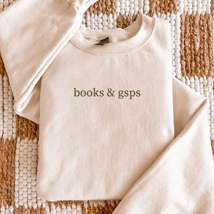 May include: A beige sweatshirt with the words "books & gsps" printed in a dark brown color. The sweatshirt is folded and placed on a patterned rug. The sweatshirt has a crew neck and long sleeves.
