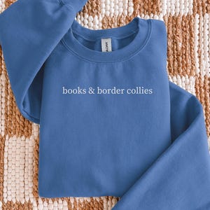 May include: A blue crewneck sweatshirt with the words "books & border collies" printed in white. The sweatshirt is folded on a patterned beige and brown surface.