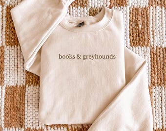 May include: A beige sweatshirt with the words "books & greyhounds" printed in a brown font. The sweatshirt is folded on a patterned rug with beige and brown tones. The sleeves are folded over the top of the sweatshirt.