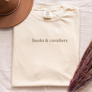 May include: Cream-colored t-shirt with the text "books & cavaliers" in brown. The shirt is folded and placed on a white surface. A brown hat and a bundle of dried purple flowers are also in the image.