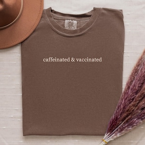 May include: A brown t-shirt with the text "caffeinated & vaccinated" in white. A brown hat and a bundle of dried, purple-toned flowers are also in the image. The shirt is made of a soft material.