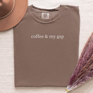 May include: A brown t-shirt with the text "coffee & my gsp" in white. The shirt is folded and lies on a white surface. A brown hat and a bundle of dried purple flowers are in the image.