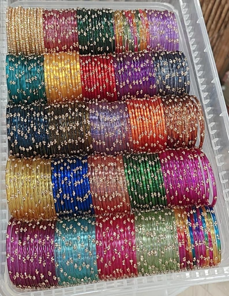 May include: A collection of colorful glass bangles in a clear plastic container. The bangles are in various colors, including gold, green, red, purple, and blue, and are decorated with small, sparkling accents. The bangles are stacked in rows.