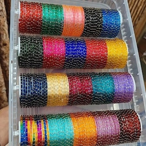 May include: A clear plastic container filled with multiple rows of colorful bangles. The bangles are in various colors, including red, green, pink, blue, yellow, and purple. Each bangle is adorned with small, reflective beads.