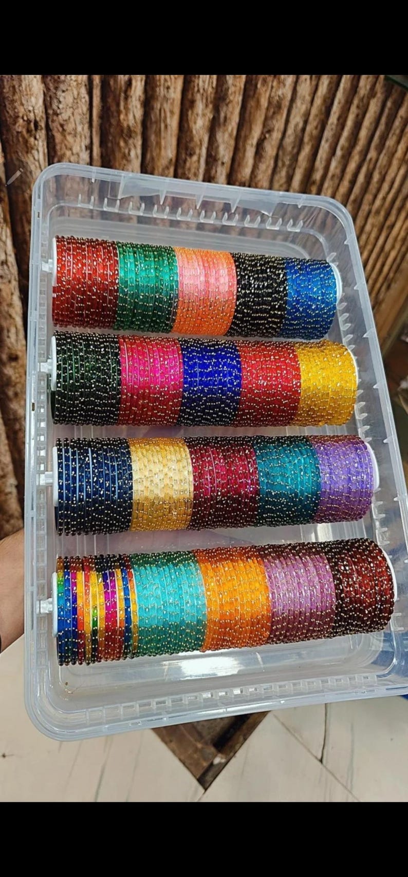 May include: A clear plastic container filled with multiple rows of colorful bangles. The bangles are in various colors, including red, green, pink, blue, yellow, and purple. Each bangle is adorned with small, reflective beads.