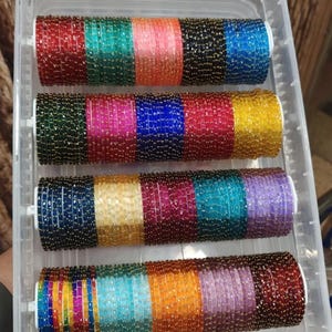 May include: A clear plastic container holding several rows of colorful bangles. The bangles are arranged in a rainbow of colors, including red, green, blue, yellow, and purple. Each bangle is adorned with small, shiny beads.