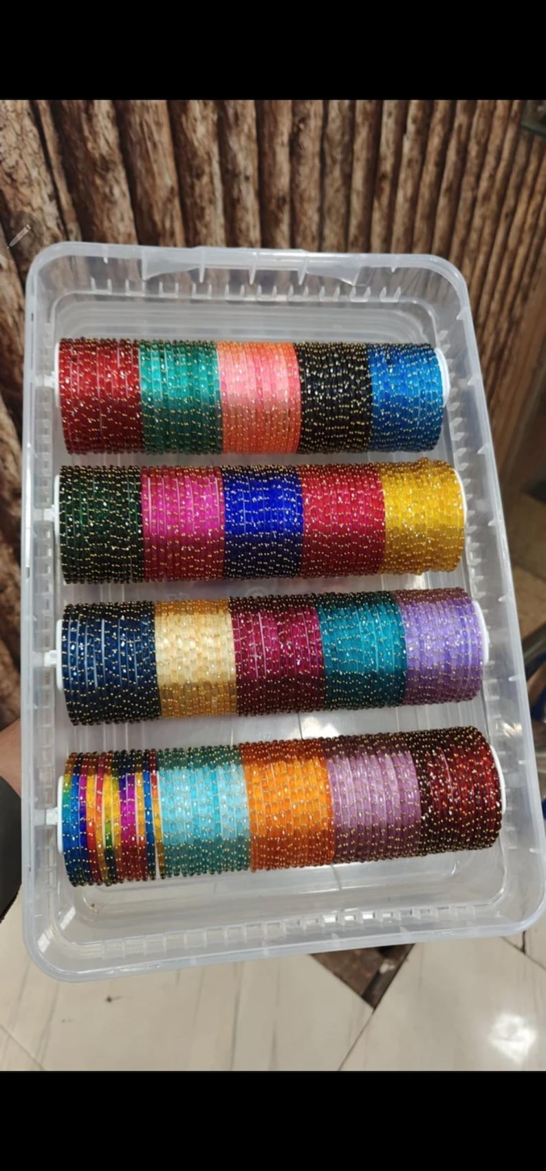 May include: A clear plastic container holding several rows of colorful bangles. The bangles are arranged in a rainbow of colors, including red, green, blue, yellow, and purple. Each bangle is adorned with small, shiny beads.