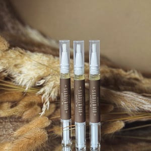 Puede incluir: Tres bolígrafos de aceite para cutículas con tapas transparentes y etiquetas marrones que dicen "+ SANDALWOOD + CUTICLE OIL PEN". Los bolígrafos están llenos de un líquido amarillo y se colocan sobre un fondo borroso de plantas secas.