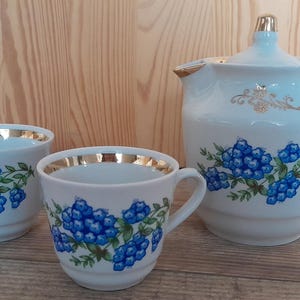 May include: A white teapot and two teacups with a blue floral pattern and gold accents. The teapot has a lid and a handle. The teacups have a handle and a gold rim.