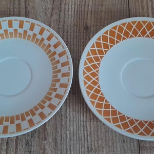 Könnte beinhalten: Zwei weiße Untertassen aus Keramik mit orangefarbenen geometrischen Mustern. Eine Untertasse hat ein Muster aus Quadraten und die andere ein Muster aus Rauten.