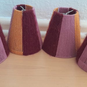 Könnte beinhalten: Vier Lampenschirme aus gewebtem Stoff in Braun-, Orange- und Lilatönen. Die Lampenschirme sind alle gleich groß und gleich geformt und in einer Reihe angeordnet.