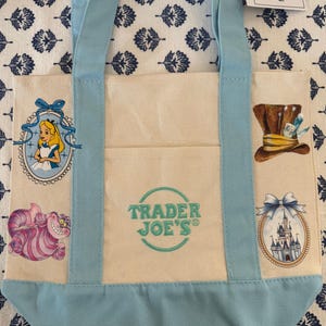 Może przedstawiać: Mini płócienna torba typu tote w pastelowych kolorach z jasnoniebieskimi akcentami i uchwytami. Zawiera ilustracje z Alicji w Krainie Czarów i logo Trader Joe's. Zawiera metkę z napisem "Mini Pastel Canvas Tote Bag".