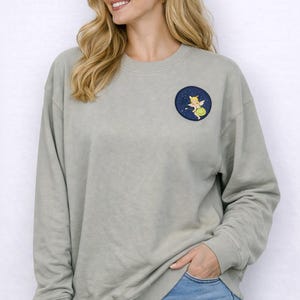 May include: A sage green sweatshirt with a round patch featuring a cartoon fairy on a navy blue background. The sweatshirt has long sleeves and is paired with blue jeans.