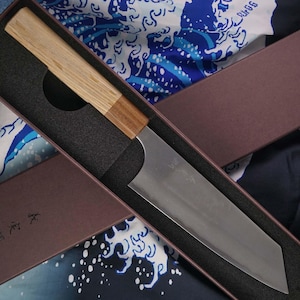 May include: A Japanese-style kitchen knife with a light wood handle and a silver blade, presented in a brown box with a dark interior. The knife has a straight edge and a slightly angled tip. The box is lined with a dark gray material.