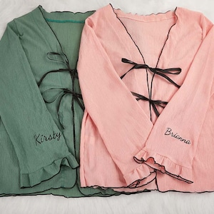 May include: Two long-sleeved robes, one sage green and one light pink, with black trim and bows. The sleeves have a ruffled edge and the names "Kirsty" and "Brianna" are embroidered on the sleeves.