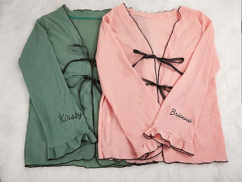 May include: Two long-sleeved robes, one sage green and one light pink, with black trim and bows. The sleeves have a ruffled edge and the names "Kirsty" and "Brianna" are embroidered on the sleeves.