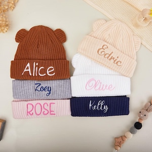 Può includere: Pila di sei berretti per bambini personalizzati in vari colori: marrone, grigio, rosa, bianco e blu navy. Ogni berretto ha un testo ricamato con nomi come "Alice", "Zoey", "Rose", "Edric", "Olive" e "Kelly". I berretti hanno orecchie da orso.