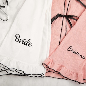 May include: White and pink robes with black trim and ties. The white robe has the word "Bride" embroidered on the sleeve, while the pink robe has the name "Brianna" embroidered on the sleeve. Both robes have ruffled edges.