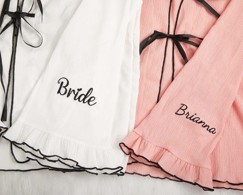 May include: White and pink robes with black trim and ties. The white robe has the word "Bride" embroidered on the sleeve, while the pink robe has the name "Brianna" embroidered on the sleeve. Both robes have ruffled edges.