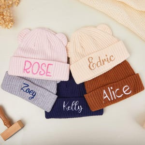 Puede incluir: Varios gorros de punto acanalado en varios colores, incluyendo rosa, gris, azul marino, crema y marrón. Cada gorro tiene nombres bordados: Rose, Zoey, Kelly, Edric y Alice. Los gorros tienen pequeños detalles de orejas.