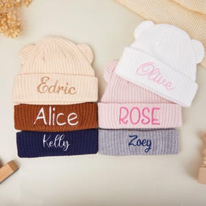 Op de afbeelding: Een stapel van zes baby mutsen in verschillende kleuren, waaronder crème, bruin, marineblauw, roze, grijs en wit. Elke muts heeft geborduurde namen zoals "Edric", "Alice", "Kelly", "Rose", "Zoey" en "Olive". De mutsen hebben een geribbelde textuur en berenoren.