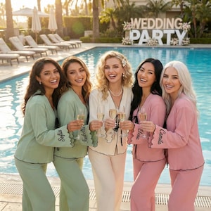 May include: Five women in matching pyjamas holding champagne flutes by a swimming pool. The pyjamas are in shades of green, pink, and cream, with black trim and personalised names on the sleeves. A sign that says "WEDDING PARTY" is in the background.