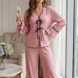 May include: A pink pajama set featuring a long-sleeved top and trousers. The top has a V-neck with black trim and a black bow. The sleeves are flared with the name "Sophia" embroidered. The set is made of textured fabric.