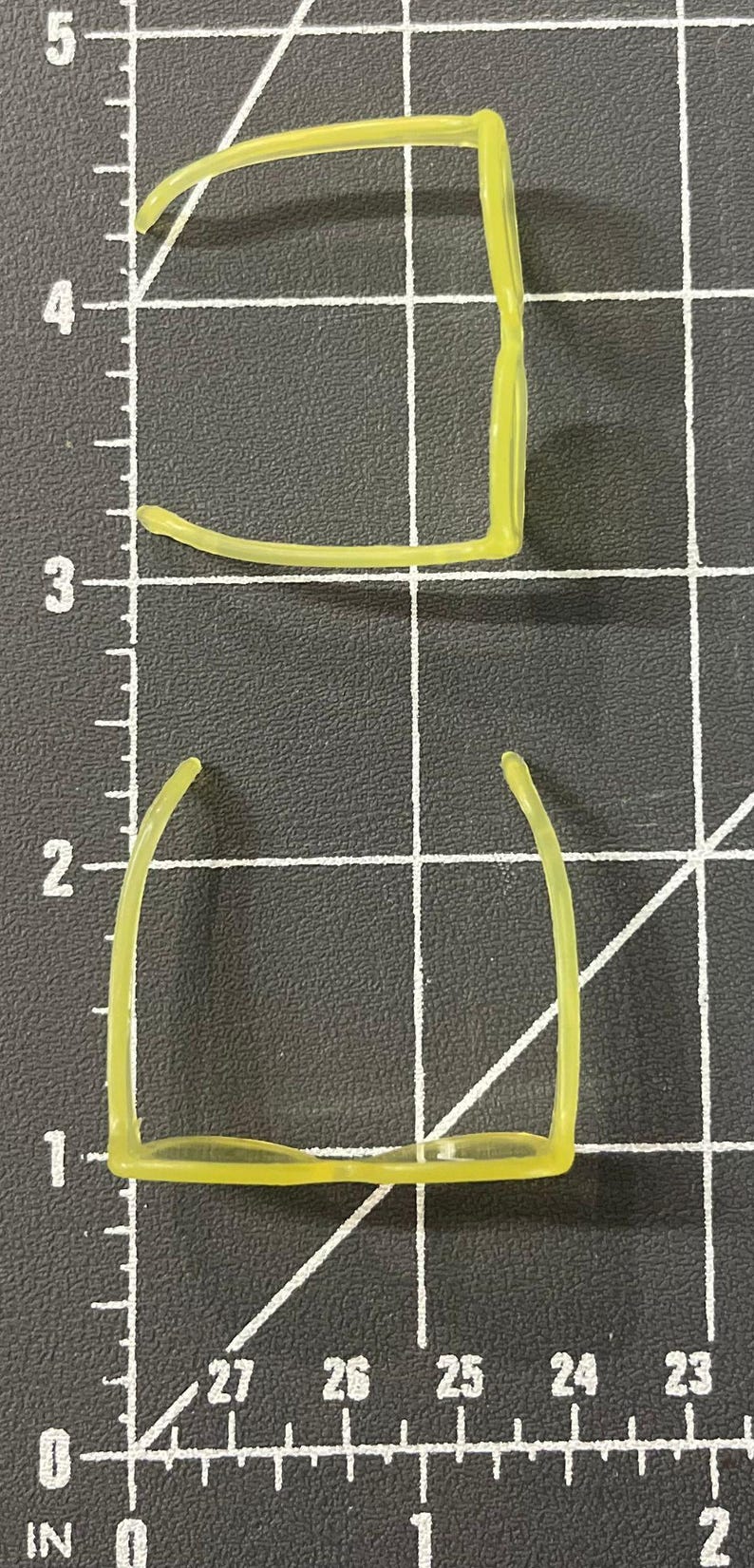 May include: Two pairs of neon yellow eyeglasses frames are displayed on a black grid surface. The frames are positioned with the top pair oriented horizontally and the bottom pair vertically. Each frame measures approximately 5 inches in length.