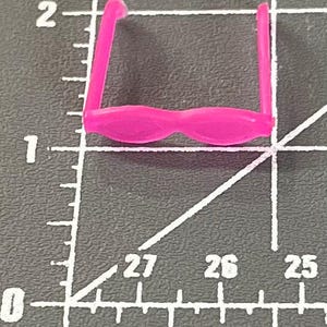 May include: A small, bright pink toy pair of glasses. The glasses have a rounded frame and straight arms. The glasses are set against a dark gray background with white measurement markings.