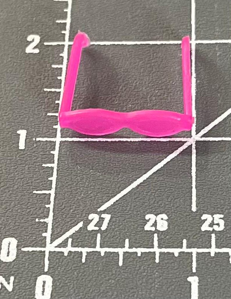 May include: A small, bright pink toy pair of glasses. The glasses have a rounded frame and straight arms. The glasses are set against a dark gray background with white measurement markings.