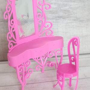 Pode incluir: Um conjunto de penteadeira de plástico rosa com um espelho e uma cadeira. A penteadeira tem um design decorativo e ornamentado com volutas e curvas. A cadeira tem um design simples com um encosto curvo e pernas.