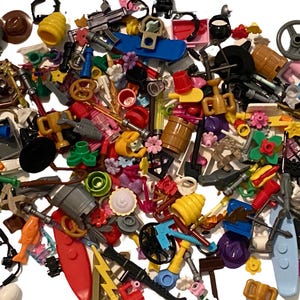 Op de afbeelding: Een grote verzameling diverse plastic bouwsteenaccessoires, waaronder miniatuurgereedschap, wapens, etenswaren, bloemen, trofeeën, vaten en diverse geometrische vormen in een breed scala aan levendige kleuren, verspreid over een wit oppervlak.
