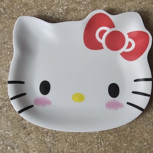 May include: White, shaped plate featuring a cartoon cat face with a red bow, black eyes, whiskers, and pink cheeks. The plate has a smooth surface and is designed with rounded edges. A fun and decorative item for serving snacks or displaying as a collectible.