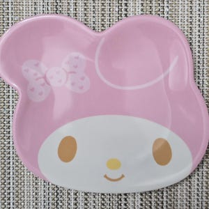 May include: A pink ceramic plate shaped like a character's head, featuring a white face, brown eyes, and a yellow nose. A bow with heart details is on the left side. The plate has a glossy finish.