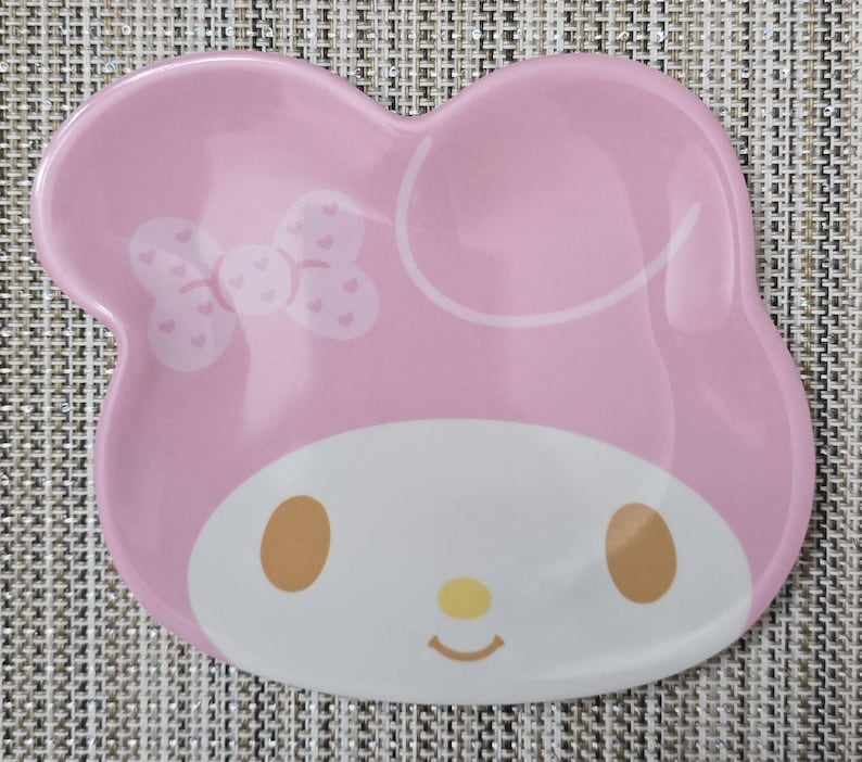 May include: A pink ceramic plate shaped like a character's head, featuring a white face, brown eyes, and a yellow nose. A bow with heart details is on the left side. The plate has a glossy finish.