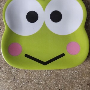 May include: A lime green ceramic tray with a cartoon frog face design. The tray features large white eyes with black pupils, pink cheeks, and a black mouth. The number "2" is visible in the top left corner.