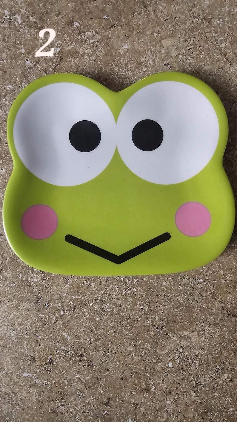 May include: A lime green ceramic tray with a cartoon frog face design. The tray features large white eyes with black pupils, pink cheeks, and a black mouth. The number "2" is visible in the top left corner.