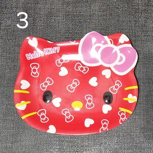 May include: A red Hello Kitty-shaped dish with a pink bow, black eyes, and yellow accents. The dish is decorated with white bows and hearts, and the words "Hello Kitty" are printed on the front. The number "3" is in the upper left corner.