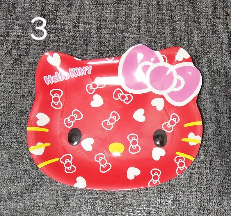 May include: A red Hello Kitty-shaped dish with a pink bow, black eyes, and yellow accents. The dish is decorated with white bows and hearts, and the words "Hello Kitty" are printed on the front. The number "3" is in the upper left corner.