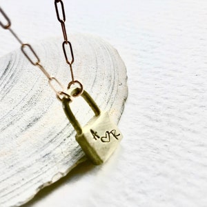 Puede incluir: Un collar de oro con un dije en forma de candado. El candado tiene las letras "K" y "R" grabadas, con un símbolo de corazón entre ellas.