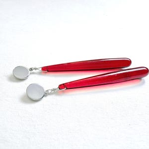 Op de afbeelding: Een paar rode druppelvormige oorbellen met zilverkleurige ronde studs. De oorbellen hebben een lang, slank en glad ontwerp. Het rode deel van de oorbellen is doorschijnend. De oorbellen worden op een wit oppervlak weergegeven.