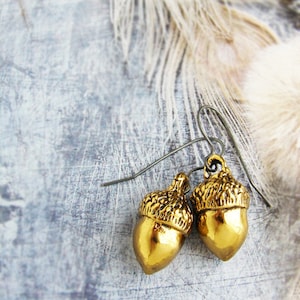 Op de afbeelding: Een paar gouden eikel oorbellen met een eenvoudig draadontwerp. De oorbellen hangen aan een witte veer op een grijze achtergrond.
