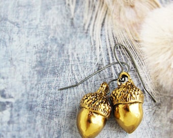 May include: A pair of gold acorn earrings with a simple wire design. The earrings are hanging from a white feather on a gray background.