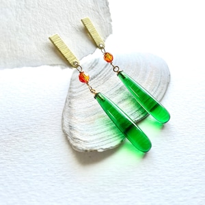May include: Dangle earrings with green glass teardrop pendants. The earrings have gold-coloured rectangular tops and orange bead accents. The earrings are displayed on a white surface.