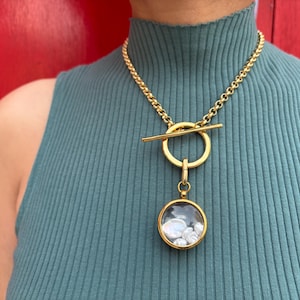 Könnte beinhalten: Eine goldene Kettenkette mit einem Toggle-Verschluss und einem runden Anhänger. Der Anhänger ist aus klarem Glas und enthält kleine weiße Perlen.