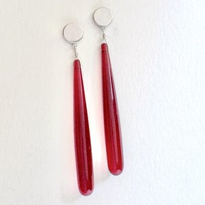 Op de afbeelding: Een paar oorhangers met lange, druppelvormige rode glazen hangers. Elke oorbel heeft een zilverkleurige ronde oorsteker en draaddetails. De oorbellen worden tegen een witte achtergrond getoond.