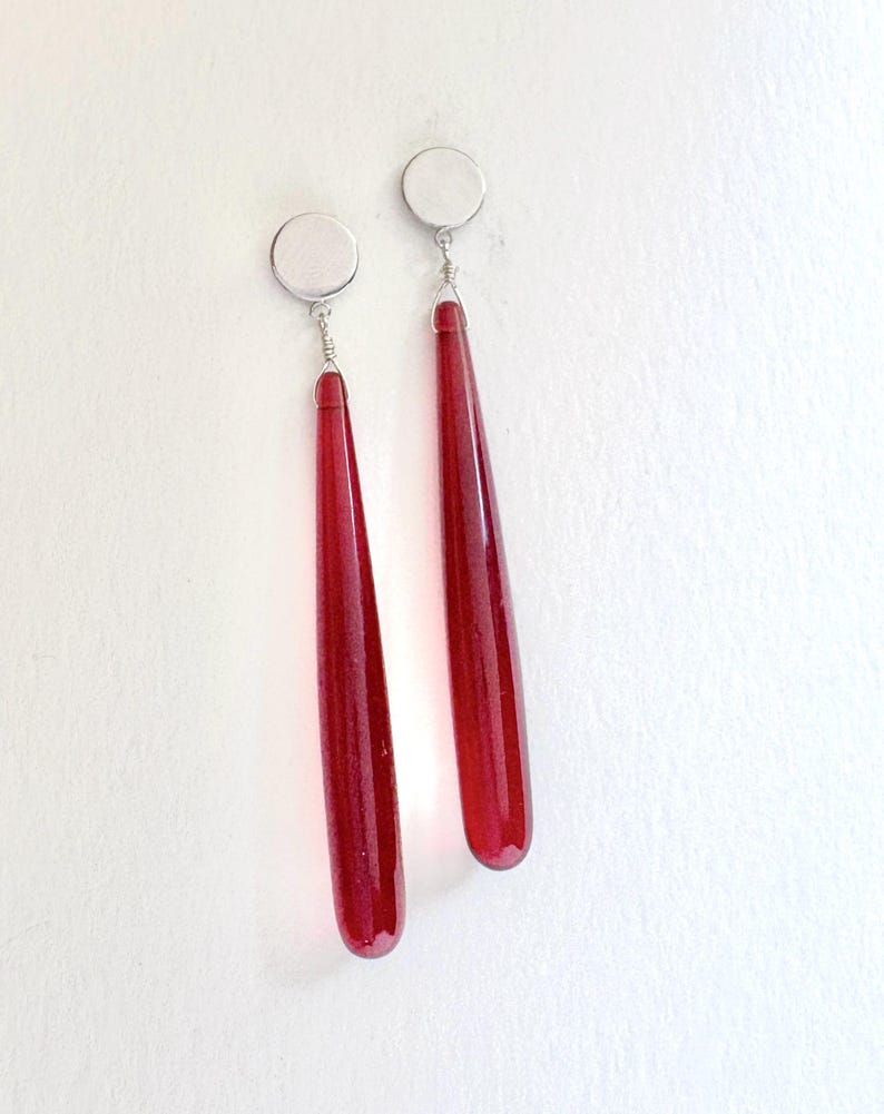 Op de afbeelding: Een paar oorhangers met lange, druppelvormige rode glazen hangers. Elke oorbel heeft een zilverkleurige ronde oorsteker en draaddetails. De oorbellen worden tegen een witte achtergrond getoond.