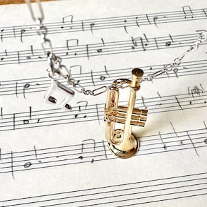 Puede incluir: Collar con colgante de trompeta dorada, cadena plateada y charm de nota musical. El collar está sobre un fondo de partituras, creando un tema musical.