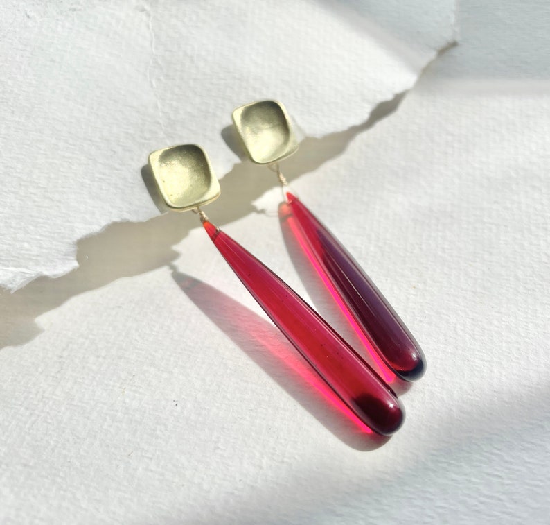 Op de afbeelding: Oorbellen met vierkante, goudkleurige studs en lange, druppelvormige hangers. De hangers zijn doorschijnend, dieprood, met een gradi&euml;nteffect. De oorbellen worden gepresenteerd op een wit, getextureerd oppervlak.