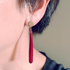 Op de afbeelding: Een gouden oorbel met een lange, druppelvormige, rode hanger. De oorbel wordt getoond op het oor van een persoon, met de rode hanger naar beneden. De oorbel heeft een eenvoudig, elegant ontwerp.
